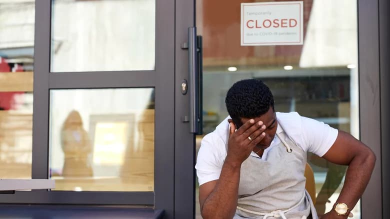 A business owner covering their face outside a shop that is temporarily closed
